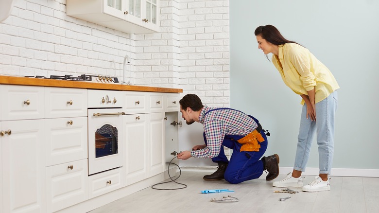 Plumber,Crouching,On,The,Floor,In,A,Modern,White,Kitchen